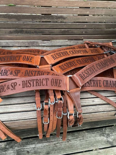 Pile of Western breast collars engraved with 'Reserve Champion' and 'District One' text on a wooden bench