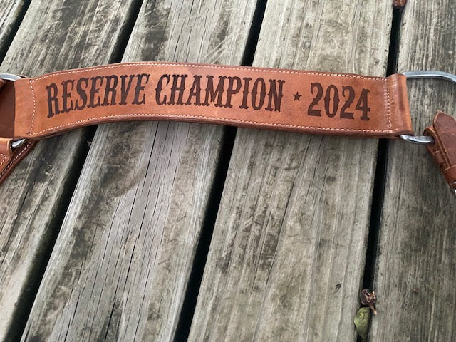 Brown leather breast collar strap with 'Reserve Champion 2024' laser engraved text on a wooden surface