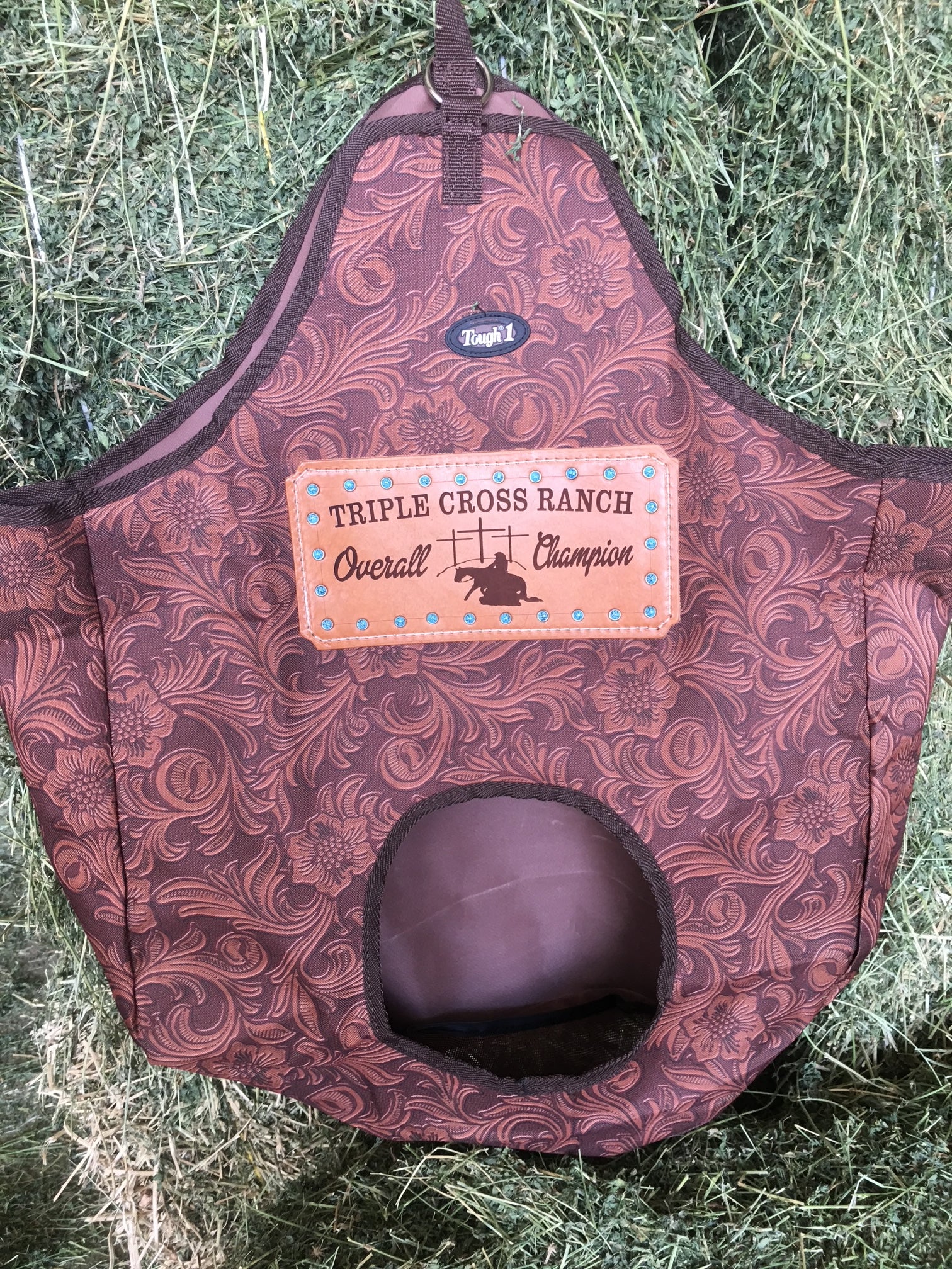 Tough 1 Brown Leather Tooled Hay Bag with a customized leather award patch for a western cowboy church, presented on a background of fresh alfalfa.