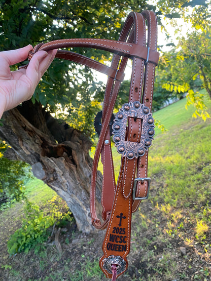 Brown leather bridle with decorative buckle and laser engraved sides held by a hand against a natural background