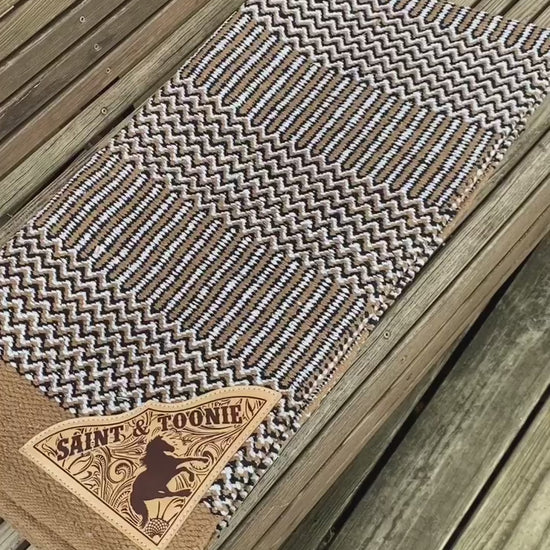 A video showing a tan navajo saddle blanket with custom corners for a horse and rider, with a leather patch laser engraved with a horse galloping over a faux leather tooling background underneath the horse and rider name.
