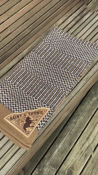 A video showing a tan navajo saddle blanket with custom corners for a horse and rider, with a leather patch laser engraved with a horse galloping over a faux leather tooling background underneath the horse and rider name.